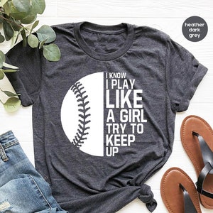 Op de afbeelding: Een donkergrijs heather t-shirt met een witte baseball-afbeelding en de tekst "I know I play like a girl try to keep up".