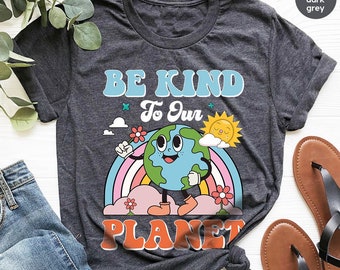 Puede incluir: Una camiseta gris oscuro jaspeada con el texto "Be Kind To Our Planet" en una fuente de estilo retro. El gráfico presenta una Tierra de dibujos animados con una cara sonriente y brazos, de pie sobre una nube con un arcoíris detrás.