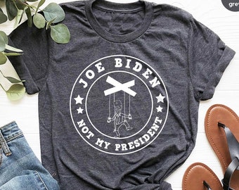 May include: Dark heather grey t-shirt with the text "JOE BIDEN NOT MY PRESIDENT" in a white circular design. The design features a puppet with strings. The shirt is paired with blue jeans and brown sandals.