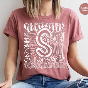 May include: A heather mauve t-shirt with a white graphic design that says "SONOGRAPHER" multiple times in a stylized font.