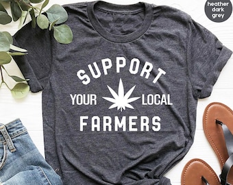 Pode incluir: Camiseta cinza escuro heather com texto branco que diz "Apoie seus agricultores locais" e um gráfico de folha de maconha.