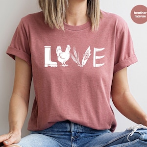 May include: A heather mauve t-shirt with a white graphic of a boot, chicken, wheat, corn, and a pitchfork spelling out the word "LOVE".