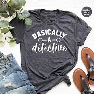 May include: Dark grey t-shirt featuring the white text "BASICALLY - A - detective". The image includes blue jeans and brown sandals. The t-shirt is a crew neck with short sleeves. The shirt is a dark heather colour.