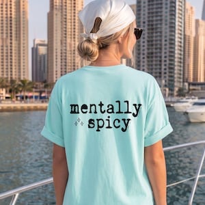 May include: A light turquoise short-sleeve t-shirt featuring the black text "mentally spicy" printed on the back, accompanied by a small four-pointed star graphic. The shirt has a crew neckline and rolled sleeves, worn with a white headscarf.