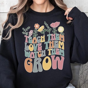 May include: Black sweatshirt with a colorful graphic that says "Teach them, love them, watch them grow" with flowers and leaves around the text.