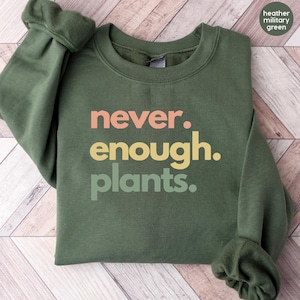 Peut inclure: Un sweat-shirt vert militaire chiné avec le texte "never. enough. plants." imprimé en orange, jaune et vert.