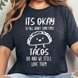May include: A woman wearing a dark grey t-shirt with the text "It's okay to fall apart sometimes Tacos do and we still love them" and a cartoon taco graphic. She is sitting on a white couch with a black and white checkered throw blanket.