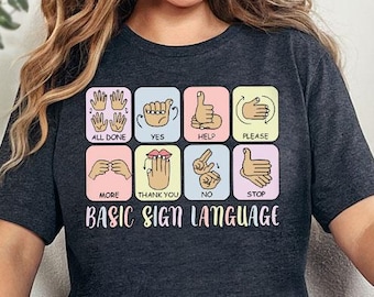 May include: A dark grey t-shirt with a graphic of basic sign language hand gestures. The graphic includes the signs for "all done", "yes", "help", "please", "more", "thank you", "no", and "stop". The text "BASIC SIGN LANGUAGE" is printed below the graphic.