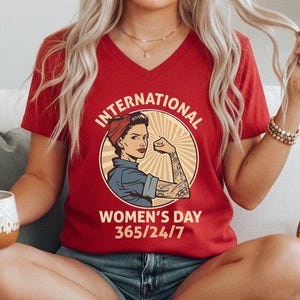 May include: Red V-neck t-shirt with a graphic of a woman flexing her arm, a red bandana, and the text "INTERNATIONAL WOMEN'S DAY 365/24/7" in a circular design. The shirt is made of a soft material.