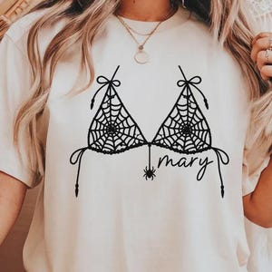 May include: A cream-coloured short-sleeve t-shirt featuring a black graphic print of a bikini top made of spider webs. A small spider hangs from the centre of the design, and the name "mary" is written in a cursive script font below the spider.