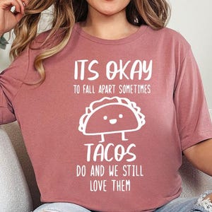 May include: A woman wearing a pink t-shirt with the text "It's okay to fall apart sometimes tacos do and we still love them" and a cartoon taco graphic. She is sitting on a white couch with a black and white checkered blanket.
