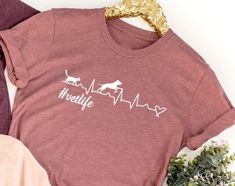 May include: Three t-shirts with a heartbeat graphic and the text #vetlife. The shirts are in different colors: maroon, pink, and a darker shade of maroon. The graphic features a cat and a dog running with a heartbeat line.