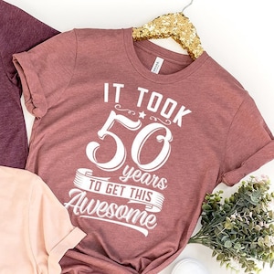 May include: Three t-shirts with the text "It took 50 years to get this Awesome" on them. The shirts are in different colors: maroon, pink, and peach.