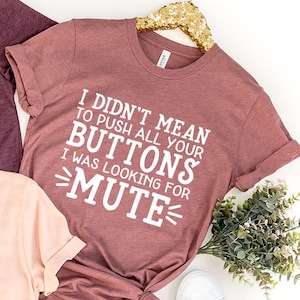 May include: Three t-shirts with the same text printed on them. The text reads "I didn't mean to push all your buttons, I was looking for mute". The shirts are in different colours: pink, maroon, and a darker maroon.