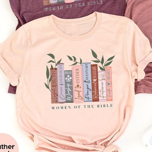 May include: Three t-shirts with a design of stacked books with names of women from the Bible. The shirts are in heather maroon, heather mauve, and heather peach.
