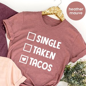 May include: Three t-shirts with a graphic design of a checkbox with the words "Single", "Taken", and "Tacos". The top t-shirt is heather maroon, the middle t-shirt is heather mauve, and the bottom t-shirt is heather peach.