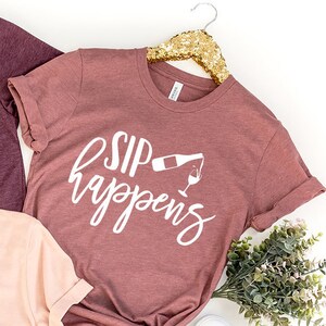 May include: Three t-shirts with the text "Sip Happens" and a graphic of a wine bottle pouring into a glass. The shirts are in different colors: maroon, light pink, and a darker shade of pink.