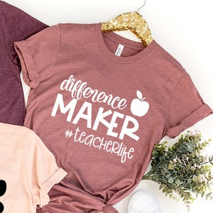 May include: Three t-shirts with the text "difference maker #teacherlife" and an apple graphic. The shirts are in pink, maroon, and a darker maroon color.