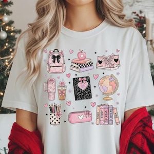 May include: A white t-shirt featuring a graphic print of school supplies including a backpack, books, sneakers, a globe, a pencil cup, and a pencil case, all decorated with pink hearts, bows, and stars, layered under a red plaid cardigan.