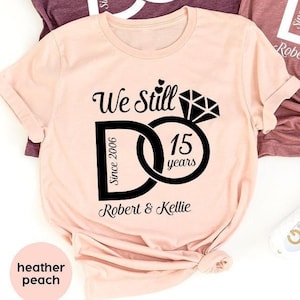 Könnte beinhalten: Drei T-Shirts mit dem Text "We Still Do" und einem Ringdesign. Die Shirts sind in den Farben Heidelbeer-Marone, Heidelbeer-Lila und Heidelbeer-Pfirsich gehalten. Der Text "Since 2006" und "15 years" ist ebenfalls auf den Shirts enthalten.