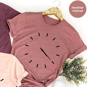 May include: Three t-shirts with a clock graphic. The shirts are in heather maroon, heather mauve, and heather peach.