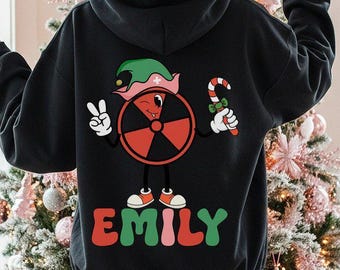 May include: A black hooded sweatshirt featuring a graphic on the back of a red radiation symbol character wearing a green elf hat, holding a candy cane, and making a peace sign. The name EMILY is printed below in green and red letters.