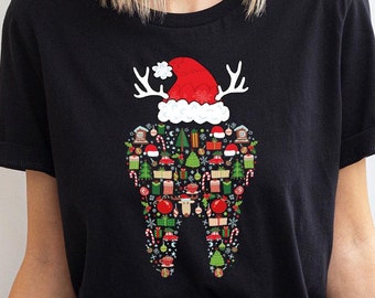 Op de afbeelding: Een zwart t-shirt met korte mouwen en een afbeelding in de vorm van een tand, gevuld met kleurrijke kersticonen zoals cadeautjes, kerstbomen, zuurstokken en kerstballen, bekroond met een rode kerstmuts en wit rendiergewei.