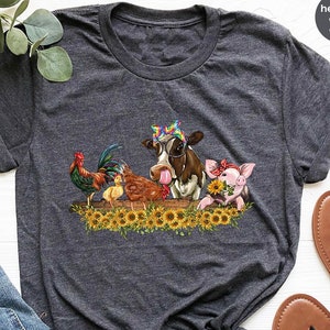 May include: A heather dark grey t-shirt with a graphic of a cow, pig, rooster, and two chicks. The animals are all wearing accessories. The cow is wearing sunglasses and a rainbow headband. The pig is wearing a red headband. The rooster is wearing a red comb. The chicks are wearing yellow beaks. The animals are all standing in front of a field of sunflowers. The t-shirt is laid out on a white surface with a pair of brown sandals.