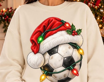 May include: A cream-coloured long-sleeve sweatshirt featuring a graphic of a football wearing a red Santa hat with white fur trim, wrapped in a string of colourful Christmas lights with green holly leaves and red berries on top.
