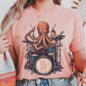 May include: A peach-colored short-sleeve t-shirt featuring a graphic illustration of a brown octopus playing a drum set. The octopus is positioned centrally on the chest, holding drumsticks and surrounded by cymbals and various drums.