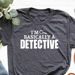 May include: A heather dark grey t-shirt with the text "I'm Basically A Detective" and a magnifying glass icon.