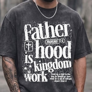 May include: Dark gray t-shirt with the text "Father is hood kingdom work" in white. The shirt also features a cross and a Bible verse. The shirt is worn with dark gray cargo shorts and a silver chain.