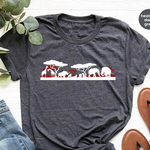 May include: Dark heather gray t-shirt featuring a white and red savanna graphic. The design includes silhouettes of giraffes, elephants, and trees. The text "heather dark grey" is in the upper right corner. The shirt is paired with blue jeans and sandals.