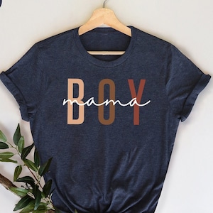 May include: A heather navy blue t-shirt with the text "mama BOY" in white and brown lettering. The letters are stacked vertically with the word "mama" in a cursive font.