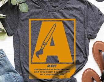 May include: A heather dark grey t-shirt with a graphic of a paintbrush inside the letter "A" in orange. The text "ART" is below the graphic with the definition "the product of imagination and creativity, particularly in a physical form."