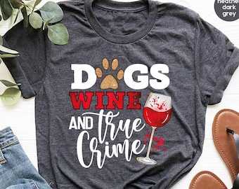 May include: Heather dark grey t-shirt with the text "Dogs, Wine, and True Crime" in white and red. The design includes a paw print, a wine glass with red wine, and the text "Fast Custom Tees". The shirt is paired with blue jeans and sandals.
