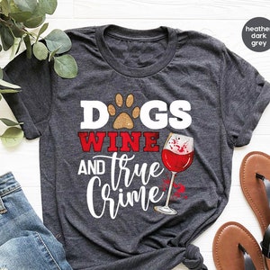 May include: Heather dark grey t-shirt with the text "Dogs, Wine, and True Crime" in white and red. The design includes a paw print, a wine glass with red wine, and the text "Fast Custom Tees". The shirt is paired with blue jeans and sandals.