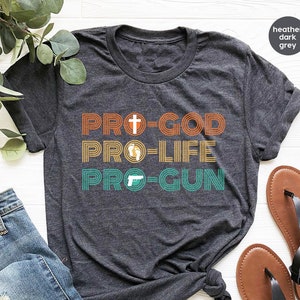 May include: Dark grey t-shirt featuring the text "PRO-GOD PRO-LIFE PRO-GUN" in orange, yellow, and teal. The design includes a cross, footprints, and a handgun. The shirt is shown with blue jeans and sandals.