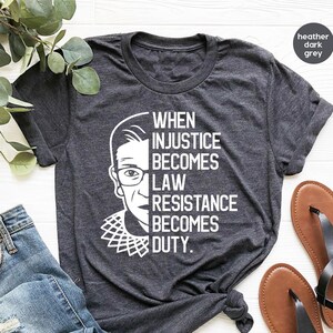 May include: A heather dark grey t-shirt with a white image of Ruth Bader Ginsburg and the text "When injustice becomes law resistance becomes duty."