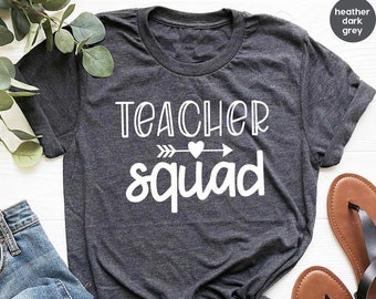 May include: A heather dark grey t-shirt with the text "teacher squad" in white. The text is accompanied by a white arrow with a heart in the middle.