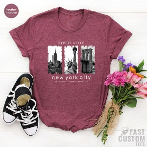 May include: Heather maroon t-shirt with a graphic print of the New York City skyline, Statue of Liberty, and Brooklyn Bridge. The text "Street Style" and "New York City" is printed on the shirt. The text "Empire State, Liberty, Brooklyn" is printed below the graphic.