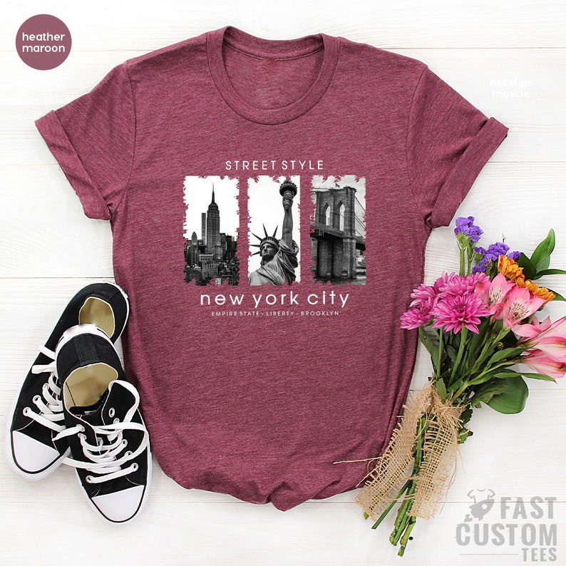 May include: Heather maroon t-shirt with a graphic print of the New York City skyline, Statue of Liberty, and Brooklyn Bridge. The text "Street Style" and "New York City" is printed on the shirt. The text "Empire State, Liberty, Brooklyn" is printed below the graphic.