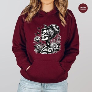 Puede incluir: Una sudadera con capucha color granate con un gr&aacute;fico blanco de una mujer esqueleto con una rosa en la mano y mariposas volando a su alrededor. El gr&aacute;fico est&aacute; rodeado de flores.