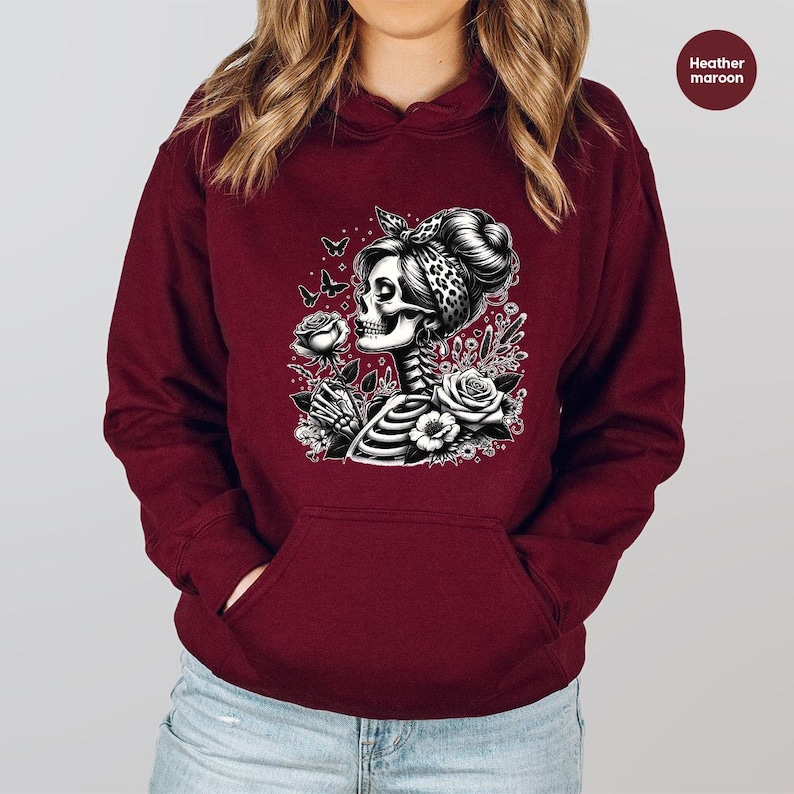 Puede incluir: Una sudadera con capucha color granate con un gr&aacute;fico blanco de una mujer esqueleto con una rosa en la mano y mariposas volando a su alrededor. El gr&aacute;fico est&aacute; rodeado de flores.