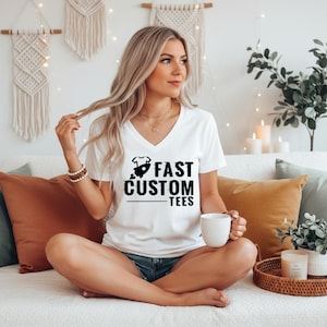 May include: White V-neck t-shirt with the text "FAST CUSTOM TEES" in black. The shirt features a graphic of a baby onesie and a rocket ship. The shirt is worn by a person sitting on a couch.
