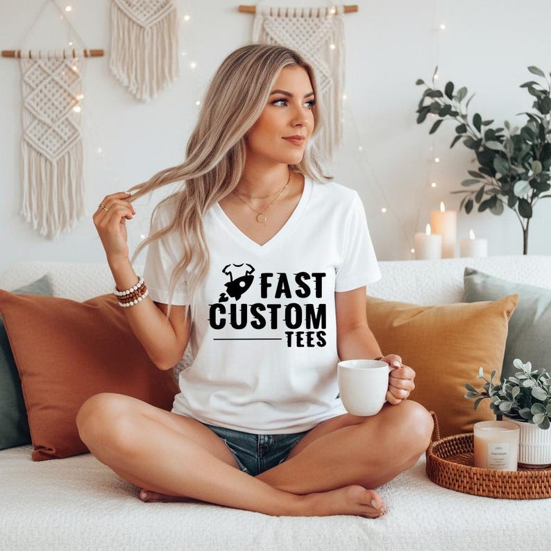May include: White V-neck t-shirt with the text "FAST CUSTOM TEES" in black. The shirt features a graphic of a baby onesie and a rocket ship. The shirt is worn by a person sitting on a couch.