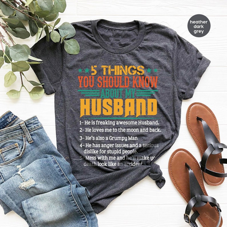 May include: A dark heather grey t-shirt with a vintage-style graphic that reads "5 Things You Should Know About My Husband". The graphic is in orange, yellow, and white. The text below the graphic lists five humorous facts about a husband.