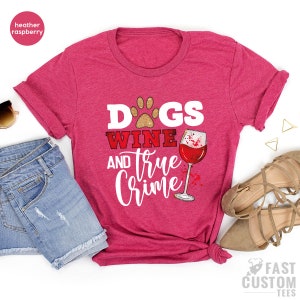 May include: Heather raspberry t-shirt with the text "Dogs Wine and True Crime" in white and red. The design includes a paw print, a wine glass, and a wine stain graphic. The shirt is paired with denim shorts and sandals.