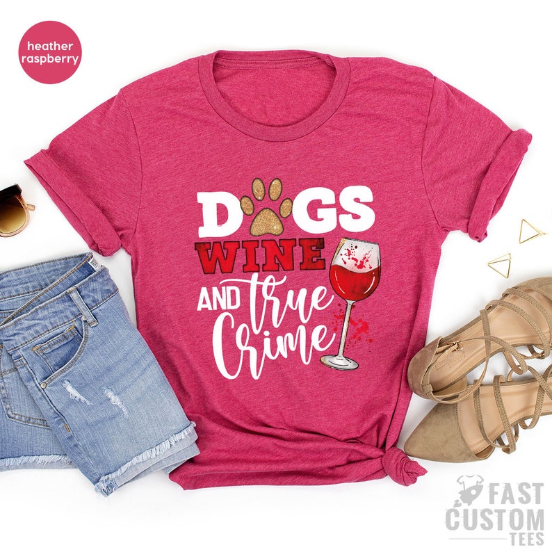 May include: Heather raspberry t-shirt with the text "Dogs Wine and True Crime" in white and red. The design includes a paw print, a wine glass, and a wine stain graphic. The shirt is paired with denim shorts and sandals.