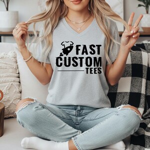 May include: A light gray V-neck t-shirt with the text "FAST CUSTOM TEES" in black. The graphic includes a rocket ship and a baby onesie. The model is wearing light blue jeans.